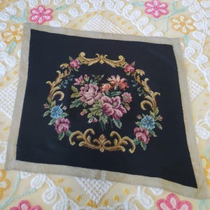 May include: A square, black needlepoint with a floral design. The central bouquet features pink, purple, and orange flowers, surrounded by a gold-colored, ornate frame. The border is a neutral beige.