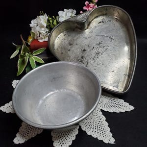 May include: Two vintage aluminum baking pans. One is a heart-shaped pan and the other is a round bowl. Both pans have a silver color and show signs of wear.