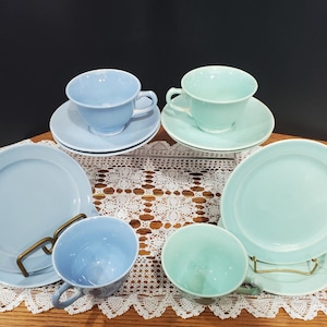 May include: A set of four teacups and saucers in two colors: light blue and light green. The cups and saucers are arranged on a white lace tablecloth.