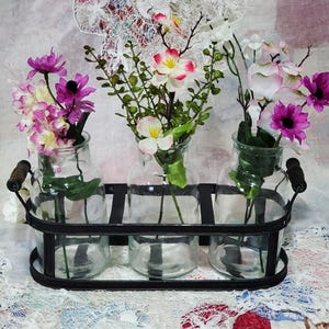 May include: A decorative floral arrangement featuring three clear glass vases filled with colorful flowers. The vases are set in a black metal tray with wooden handles. The flowers include shades of pink, purple, and white.