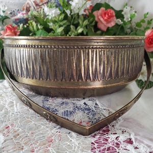 May include: A round brass bowl featuring a vertical fluted pattern along the exterior sides, a beaded rim detail, and a hinged metal handle decorated with small embossed floral motifs. The metal surface displays a warm, aged patina throughout.