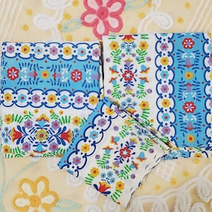 May include: Three pieces of white fabric featuring a repeating pattern of blue scalloped borders, yellow, red, and purple flowers, and green leafy vines. The design includes vertical stripes of floral motifs and decorative scalloped bands.