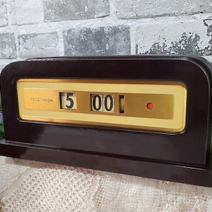 May include: A vintage brown Telechron clock displaying the time as 5:00. The clock has a rectangular shape with a gold-colored face and a red dot. The word "TELECHRON" is printed on the face.