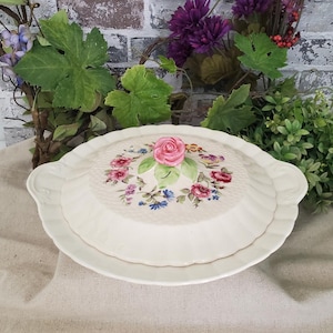 May include: Cream-colored ceramic serving dish with a lid, featuring a raised floral design with a pink rose centerpiece and accents of red, blue, and purple flowers. The dish has a textured surface and scalloped edges.