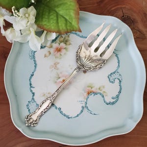Peut inclure: Une fourchette de service en argent ornée avec un manche décoratif, reposant sur une assiette à motif floral bleu et blanc.