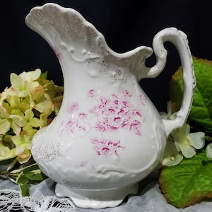 May include: White ceramic pitcher with a ruffled rim and a curved handle. The pitcher is decorated with pink floral patterns and accents. The rim and base have a textured, speckled design. A decorative piece for the home.