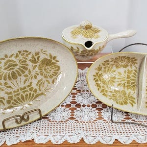 May include: A set of three white ceramic serving dishes with a green floral pattern. The dishes include a large oval platter, a smaller oval dish with a divider, and a small round dish with a lid and a handle.