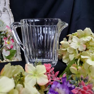 Può includere: Una lattiera in vetro trasparente e scanalato con manico e beccuccio. La lattiera è circondata da fiori artificiali colorati nelle tonalità del rosa, giallo e viola. Lo sfondo è un tessuto scuro.