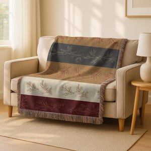 May include: A woven throw blanket draped over a beige sofa. The blanket features horizontal stripes in shades of brown, gray, cream, and burgundy, with a fringed edge. Delicate botanical patterns adorn the stripes, creating a rustic, cozy aesthetic.
