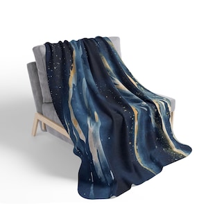 Może przedstawiać: Granatowy koc narzucony na szary fotel. Koc ma akwarelowy wzór z płynnymi niebieskimi, złotymi i białymi akcentami. Wzór koca ma motyw niebieski lub abstrakcyjny.