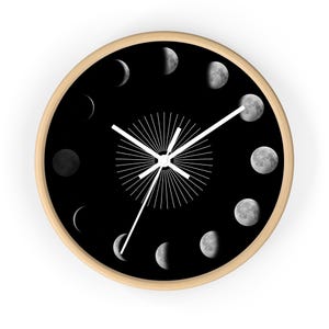 Pode incluir: Relógio de parede redondo com moldura de madeira clara e mostrador preto. O mostrador do relógio apresenta as fases da lua em um padrão circular. Ponteiros brancos e um design de explosão solar branco no centro completam o design.