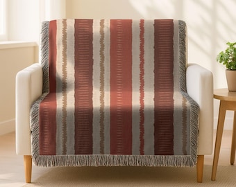 Puede incluir: Una manta con un patrón de rayas verticales en tonos rojos, beige y grises, colocada sobre un sillón blanco. La manta tiene un borde con flecos. La habitación tiene una paleta de colores neutros con una pequeña planta en maceta sobre una mesa auxiliar.