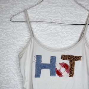 May include: White tank top with the word "HOT" spelled out in different fabrics. The "H" is made of blue denim, the "O" is made of red and white gingham, and the "T" is made of leopard print fabric.