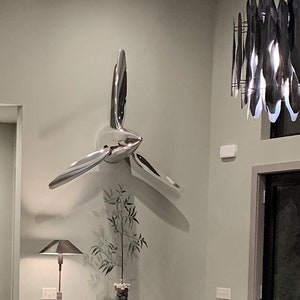May include: A silver airplane propeller sculpture mounted on a light green wall with a dark wood console table underneath. The console table has a small potted plant on top. A modern light fixture hangs from the ceiling.