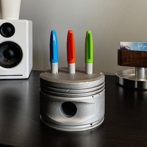 May include: A silver metal desk organiser shaped like a car engine piston with three slots for pens. Three pens are in the slots: one blue, one orange, and one green. The organiser is on a dark wood surface.