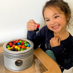 Puede incluir: Un recipiente de metal plateado con una abertura redonda y un borde elevado. El recipiente está lleno de caramelos coloridos, incluyendo M&Ms rojos, amarillos, verdes y marrones. Una niña sostiene un M&M rojo en su mano.