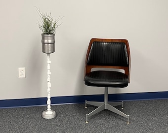 May include: A black office chair with a wooden frame and a silver base sits on a gray carpet. A plant in a silver pot sits on a white stand.