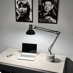 Puede incluir: Una lámpara de escritorio negra con un brazo flexible y un escritorio blanco con un ordenador portátil encima. La lámpara está encendida y proyecta luz sobre el escritorio. Hay dos fotos enmarcadas en blanco y negro en la pared detrás del escritorio.