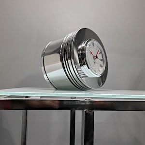 Puede incluir: Un reloj plateado con una esfera blanca y marcadores de hora romanos negros. El reloj tiene forma de cilindro y tiene una banda plateada alrededor del medio. El reloj está sentado sobre una superficie de vidrio.