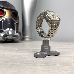 Könnte beinhalten: Eine silberne Uhr mit weißem Zifferblatt und silbernem Metallband wird auf einem grauen Metalluhrenständer präsentiert.