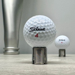 May include: A white golf ball with the text "Titleist 4" printed on it. The golf ball is sitting on a silver metal stand.