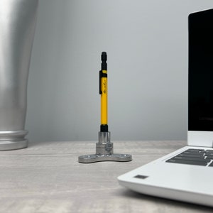 Może przedstawiać: Żółty ołówek mechaniczny z czarną gumką i czarną klamrą stoi prosto na srebrnym metalowym stojaku z trzema nogami. Stojak stoi na szarej powierzchni.