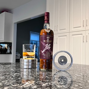 May include: A bottle of The Macallan Highland Single Malt Scotch Whisky with a red label and a gold foil top. The bottle is sitting on a counter next to a glass of whiskey and a silver bottle stopper.