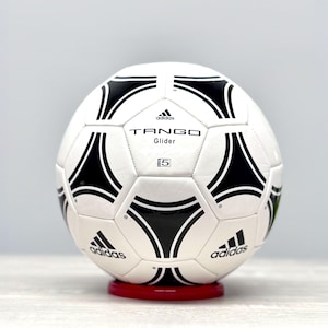 Może przedstawiać: Piłka nożna Adidas Tango Glider w kolorze białym i czarnym, ze szwami w kolorze czarnym i logo Adidas. Na piłce widnieje napis "TANGO Glider" i "5".