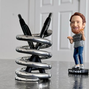 Puede incluir: Un portalápices cromado con diseño en espiral, que sostiene dos bolígrafos negros. Cerca hay una figura de cabeza oscilante de un hombre con barba y camiseta gris. La base de la figura dice "PORTNOY."