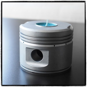 May include: A silver metal candle holder shaped like a car engine piston with a teal blue candle inside.
