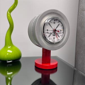 Puede incluir: Un reloj plateado con una base roja y un diseño de hélice en la esfera. El reloj está hecho para parecerse a un motor de avión.