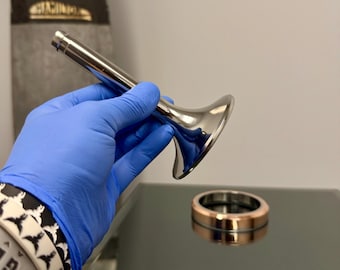 May include: A shiny, silver metal object with a flared base and a long, cylindrical handle is held in a blue-gloved hand. A gold and silver ring sits on a reflective surface. The background is a light gray.