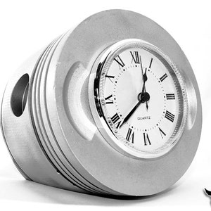 May include: A silver clock with Roman numeral markings on the face, set inside a circular frame made from a repurposed engine piston. The clock face reads "QUARTZ".