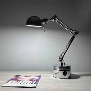 Puede incluir: Una lámpara de escritorio negra y plateada con un brazo ajustable y una pantalla redonda negra. La base de la lámpara es un pistón de metal pulido. Una revista con el título "MIDJOURNEY" está sobre la mesa.