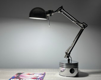 May include: A black and silver desk lamp with an adjustable arm and a round, black lampshade. The base of the lamp is a polished metal piston. A magazine with the title "MIDJOURNEY" is on the table.
