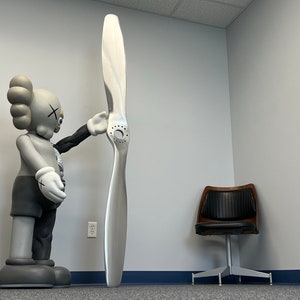 May include: A large gray and black figurine of a cartoon character holding a large silver airplane propeller. The figurine is standing on a gray carpet in a room with white walls and a black leather chair.