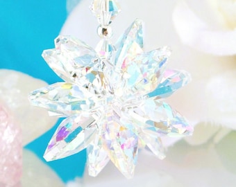 Puede incluir: Un adorno de cristal transparente y multifacetado con forma de estrella o flor, que presenta reflejos de luz iridiscentes. El adorno cuelga de un hilo de cuentas de cristal más pequeñas, transparentes y con forma de rombo.