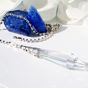 May include: A clear crystal pendulum with a silver chain and a silver ornate top. The pendulum is hanging from a chain of silver beads. The chain is resting on two blue crystals.