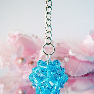 May include: A light blue crystal pendant hangs from a silver chain. The pendant is shaped like a sphere and is made up of many small, faceted crystals. The pendant is reflected in a shiny surface.