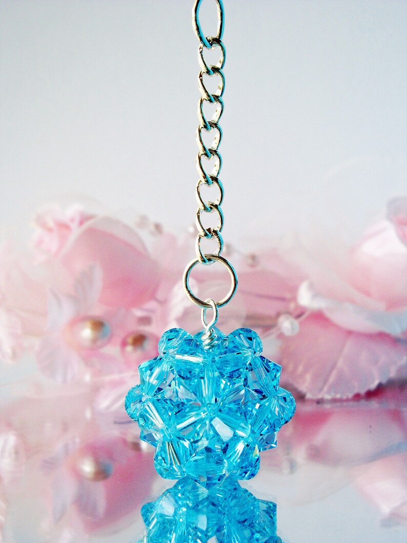 May include: A light blue crystal pendant hangs from a silver chain. The pendant is shaped like a sphere and is made up of many small, faceted crystals. The pendant is reflected in a shiny surface.