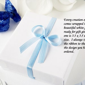 Puede incluir: Una caja de regalo blanca con una cinta azul claro atada en un lazo. La caja mide 8,9 cm por 8,9 cm por 2,5 cm. El texto en la caja dice: "Cada creaci&oacute;n m&iacute;a viene envuelta en una hermosa caja de regalo blanca lista para regalar. Esta mide 8,9 cm x 8,9 cm x 2,5 cm. Siempre intento hacer coincidir la cinta con el color del dise&ntilde;o que has pedido."