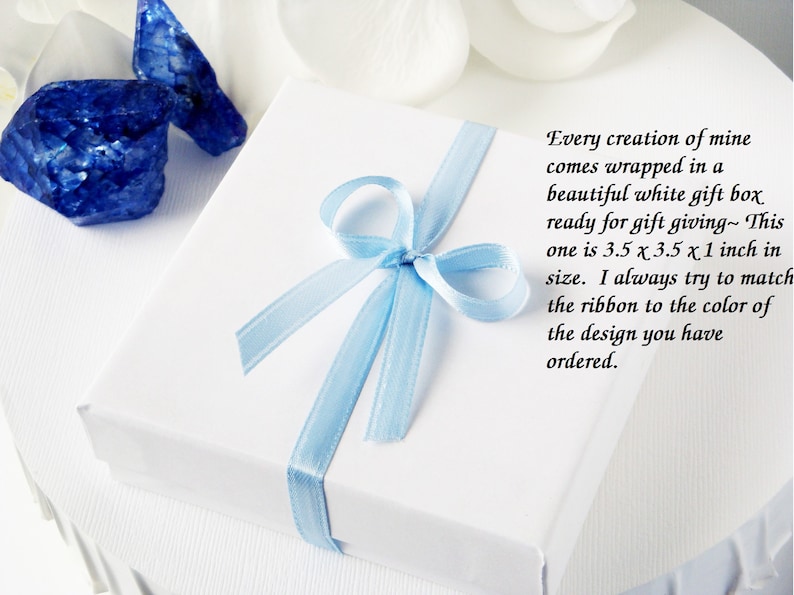 Puede incluir: Una caja de regalo blanca con una cinta azul claro atada en un lazo. La caja mide 8,9 cm por 8,9 cm por 2,5 cm. El texto en la caja dice: "Cada creaci&oacute;n m&iacute;a viene envuelta en una hermosa caja de regalo blanca lista para regalar. Esta mide 8,9 cm x 8,9 cm x 2,5 cm. Siempre intento hacer coincidir la cinta con el color del dise&ntilde;o que has pedido."