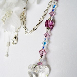 Puede incluir: Un atrapasol de cristal en forma de coraz&oacute;n con una cadena plateada y cuentas de cristal rosa y transparente. El atrapasol est&aacute; colgado de una flor de tela blanca.