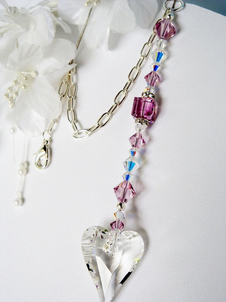 Puede incluir: Un atrapasol de cristal en forma de coraz&oacute;n con una cadena plateada y cuentas de cristal rosa y transparente. El atrapasol est&aacute; colgado de una flor de tela blanca.