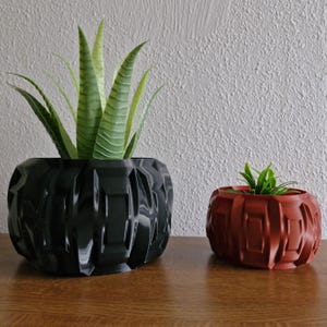 Peut inclure: Deux pots de fleurs à motifs géométriques, l'un noir et l'autre rouge, avec des plantes vertes à l'intérieur. Le pot noir est plus grand et contient une plante grasse, tandis que le pot rouge est plus petit et contient une petite plante verte.