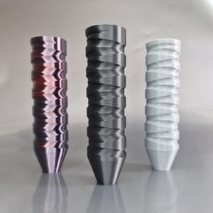 Könnte beinhalten: Drei zylindrische Dekorationsgegenstände mit einem strukturierten, wellenartigen Design. Die Objekte sind in den Farbtönen Rot-Violett, Dunkelgrau und Weiß gehalten. Jeder hat einen konischen Sockel und eine zylindrische Oberseite.
