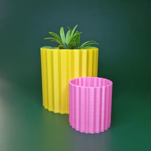May include: Two ribbed, cylindrical planters. One is bright yellow and holds a green plant. The other is smaller and pink. Both planters have a ridged design and sit on a green surface. The planters are empty.
