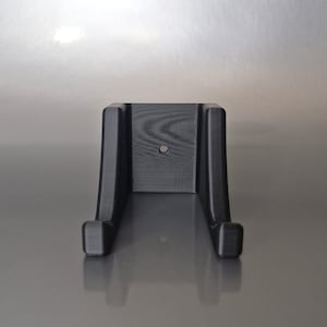 Peut inclure: Support mural noir imprimé en 3D, avec un dos rectangulaire et deux bras incurvés s'étendant vers le bas. Un petit trou est centré sur le dos. Le support est conçu pour maintenir un objet.