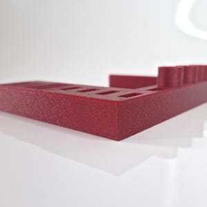May include: A burgundy-colored, rectangular desk organizer with multiple slots and cylindrical holders. The organizer is made of a textured material and is designed to hold pens, pencils, and other office supplies. The design is modern and minimalist.