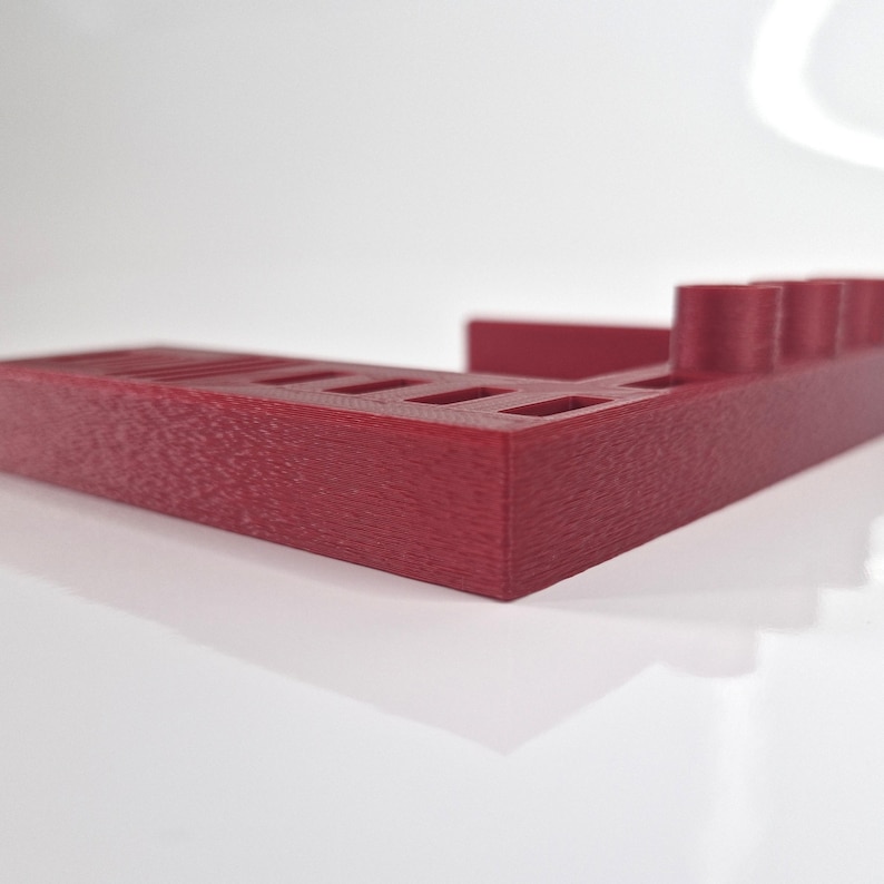 May include: A burgundy-colored, rectangular desk organizer with multiple slots and cylindrical holders. The organizer is made of a textured material and is designed to hold pens, pencils, and other office supplies. The design is modern and minimalist.